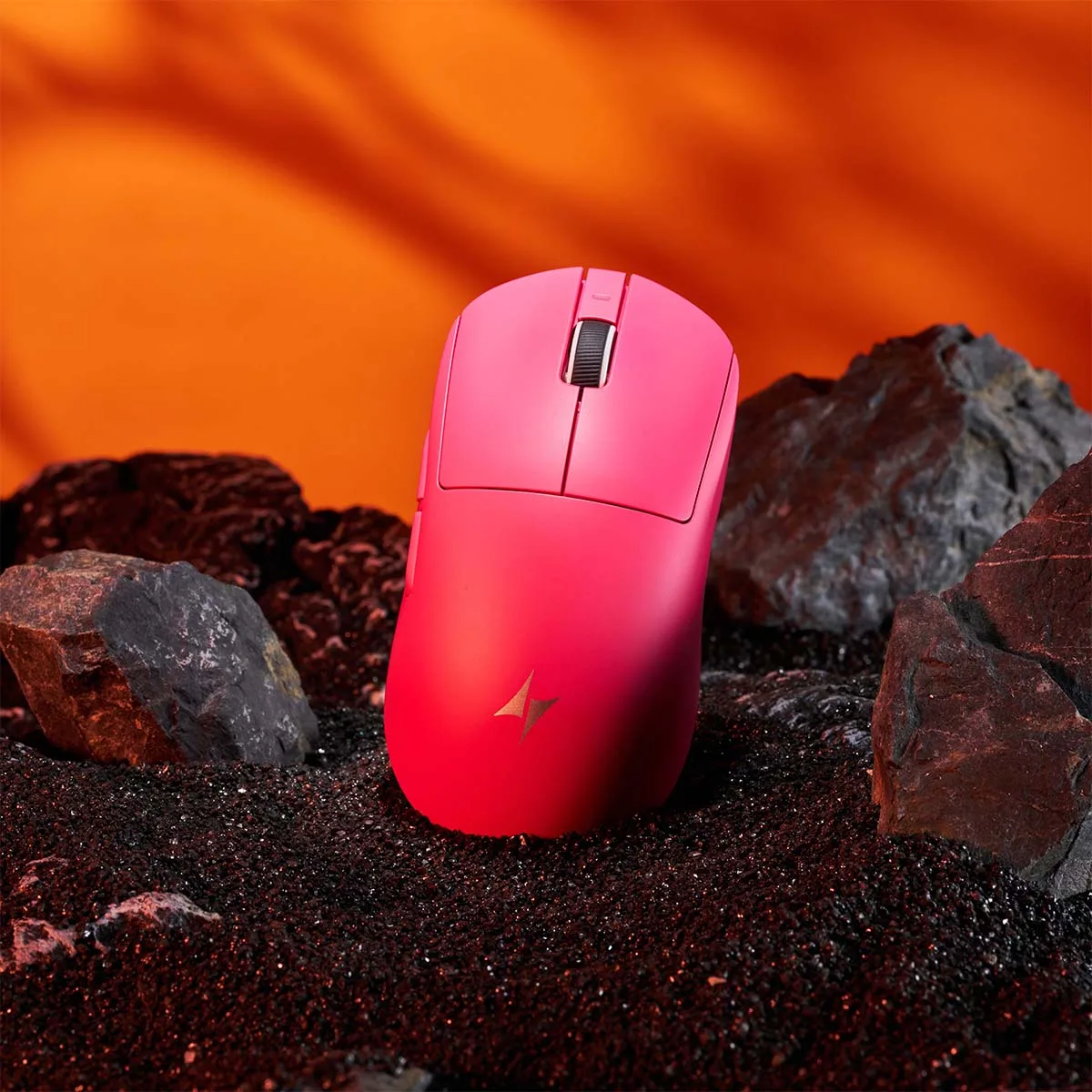 Pink ATK Dragonfly A9 gaming mouse on black sand with rocks. Dramatic orange background.